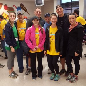 A group of eight people, some wearing yellow event T-shirts, pose and smile indoors at what appears to be a community event or race. Others wear jackets or athletic clothes. Colorful murals decorate the background wall.
