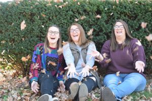 Three women sitting on the ground in front of a bush, laughing and tossing autumn leaves into the air, enjoying a playful moment together.