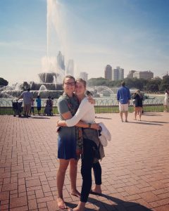 Two women smile and hug each other in front of a large fountain in a city park, with people walking around and tall buildings visible in the background on a sunny day.