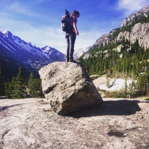 A person wearing a backpack stands on top of a large boulder, surrounded by mountains, pine trees, and patches of snow under a blue sky in a scenic wilderness setting.