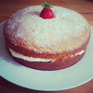 A round sponge cake with a layer of cream filling, dusted with powdered sugar on top, and decorated with a single strawberry, served on a white plate.