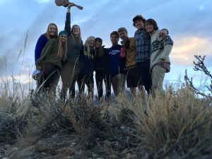 A group of nine young people standing closely together outdoors in tall grass, smiling at the camera. One person is holding a ukulele above their head. The sky is cloudy and the sun is low.