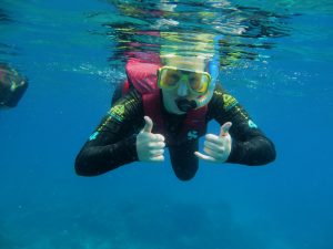 A person wearing a snorkel mask, yellow goggles, and a red life vest gives two thumbs-up while swimming underwater in clear blue sea.