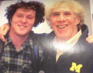 Two smiling men stand close together. The younger man on the left has curly hair and wears a plaid shirt, while the older man on the right has gray hair, glasses, and a dark jacket with a yellow M on it.
