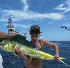 A smiling woman in sunglasses and a cap holds a large yellow and green fish on a boat, with an offshore oil platform and blue sky in the background.