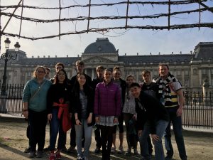 A group of twelve young people pose and smile in front of a historic building with a dome and ornate architecture, under a leafless wooden trellis on a sunny day.