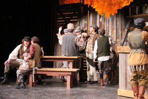 A group of actors in period costumes gather around wooden tables and crates on stage, with some standing, talking, and gesturing, set against a rustic wooden backdrop and warm lighting.