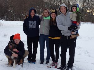 Six people and a dog pose together outdoors in the snow. Some wear bright hats and hoodies. One person holds a small child in a green coat. Trees with bare branches are visible in the snowy background.