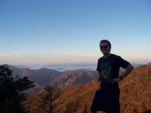 A person wearing sunglasses stands on a mountain with hands on hips, smiling at the camera. Mountains with autumn-colored trees and a blue sky stretch out in the background.