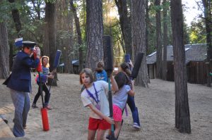 Children and an adult in a forested area playfully pose with foam bats, surrounded by tall trees and cabins in the background. Sunlight filters through the trees, creating a warm atmosphere.