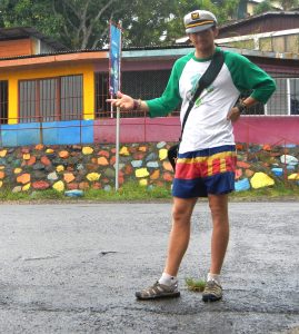 A person wearing a captains hat, green and white shirt, colorful shorts, and sandals stands on a wet street, gesturing with one hand. Behind them is a building with a colorful stone wall and bright yellow and orange paint.