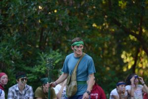 A man with green face paint, wearing a blue shirt and a green headband, holds a torch and stands outdoors. Several people, also with face paint and headbands, sit in the background among trees.
