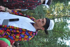 A man wearing a colorful, patterned jacket over a white shirt, with a bandana and chefs hat, stands outdoors in a wooded area, looking serious; people and trees are visible in the background.