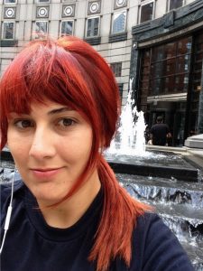 A person with red hair and a slight smile takes a selfie in front of a modern building and a tiered water fountain. They are wearing a dark shirt and white earphones.