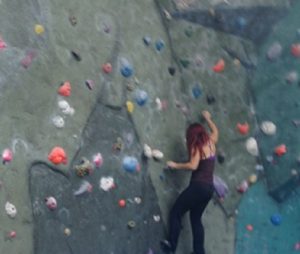 A person with red hair climbs an indoor rock climbing wall, reaching for a hold with one hand while gripping the wall with the other. They are wearing a dark tank top and pants. Colorful holds are scattered across the wall.