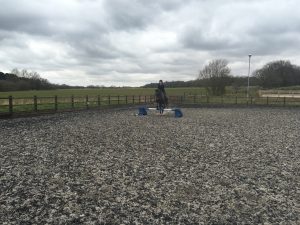 A person rides a horse in an outdoor riding arena, jumping over a small obstacle with blue supports, surrounded by wooden fencing and fields under a cloudy sky.