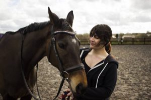 A woman with dark hair in a ponytail stands next to a brown horse in an outdoor riding arena, gently holding the horse’s reins and smiling while the wind blows her hair.