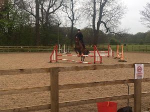 A person riding a horse jumps over a red and white hurdle in an outdoor sandy equestrian arena, surrounded by a wooden fence and trees in the background.