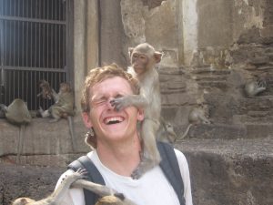 A young man laughs as a monkey playfully covers his eyes with its paw. Other monkeys sit on ancient stone ruins in the background. The man is wearing a white shirt and a backpack.