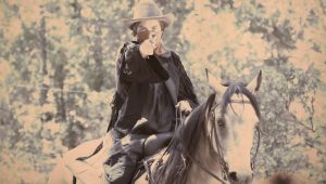 A person in a cowboy hat and dark clothing rides a horse in a wooded area, pointing a finger like a gun toward the camera. The image has a vintage, sepia-toned effect.