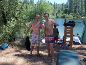 Two men in swim trunks, smiling and giving a thumbs-up, stand by a lake with trees in the background. They are covered in dirt or grass, and others are seen relaxing near the water and a wooden structure.