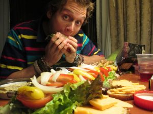 A man in a colorful striped shirt is eating a large sandwich at a table covered with sliced cheese, onions, tomatoes, lettuce, crackers, and other sandwich ingredients.