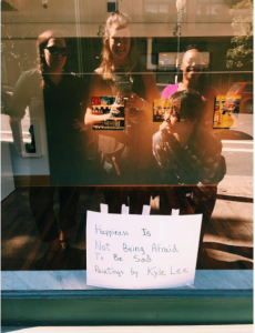 Four people are reflected in a shop window, smiling as they take a photo. Inside the window is a handwritten sign that reads, Happiness is not being afraid to be sad. Paintings by Kyle Lee.
