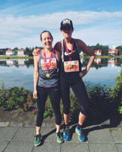 Two women wearing athletic clothes and race bibs stand side by side, smiling with medals around their necks. They are outdoors near a lake, with houses and greenery in the background on a sunny day.