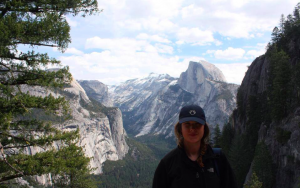 A person wearing a dark jacket and cap stands in front of a scenic mountain view, with pine trees and dramatic cliffs under a partly cloudy sky, likely in Yosemite National Park.