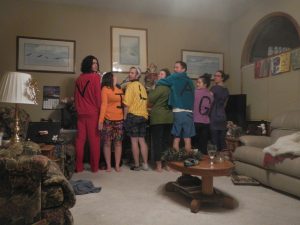 Eight people stand indoors with their backs to the camera, each wearing a brightly colored shirt with a large letter on it. Together, the shirts spell out VILLAGE. The room is warmly lit and cozy with family-style decor.