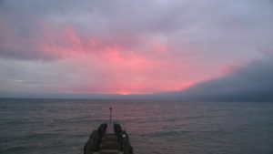A wooden pier extends into a calm sea under a cloudy sky. The horizon glows with pink and orange hues from the setting sun, casting a soft light on the water and clouds.