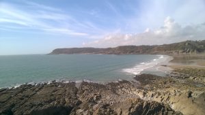 Rocky coastline with gentle waves lapping against the shore, a sandy beach curving along the bay, and distant cliffs under a partly cloudy sky.