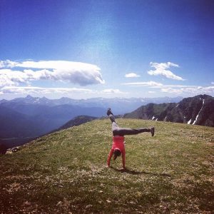 A person does a cartwheel on a grassy mountain ridge under a bright blue sky with scattered clouds, surrounded by distant mountains.