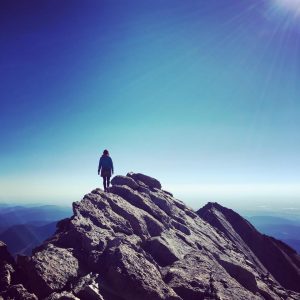 A person stands on a rocky mountain peak under a clear blue sky with the sun shining brightly, overlooking distant hills and valleys.