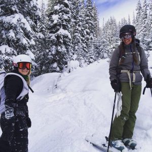 Two people wearing ski gear stand on a snowy slope surrounded by snow-covered trees. Both are smiling and appear to be enjoying their time in the winter landscape.