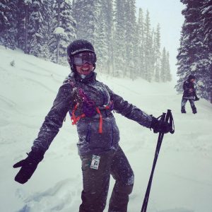 A person in winter gear smiles and poses with ski poles on a snowy slope surrounded by trees, while snow falls and another person stands in the background.