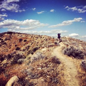 A hiker with a backpack walks along a narrow dirt trail on a dry, grassy hill under a blue sky with scattered clouds. The landscape is rugged with shrubs and rocks.