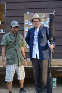 Two men stand outside in front of a bulletin board. One wears a green shirt, shorts, and a cap, while the other wears a hat, blue blazer, and holds a large black prop. Both have whistles around their necks.