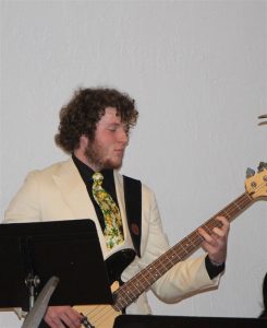 A young man with curly hair, wearing a cream-colored suit and a floral tie, plays an electric bass guitar in front of a white wall. Music stands are visible in the foreground.