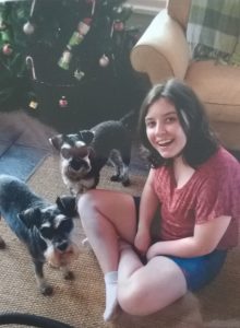 A smiling young woman sits on the floor near a decorated Christmas tree with two small black and gray dogs, one sitting beside her and the other standing behind her.