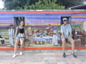 A young woman and an older man pose by sticking their heads through holes in a colorful mural of a lively outdoor market scene, with baskets of fruit and people painted on tiled walls behind them.
