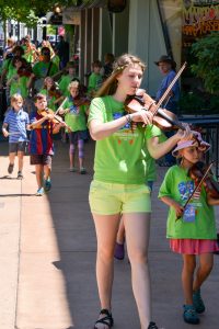A group of children and teens, all wearing bright green T-shirts, play violins while walking down a sunny sidewalk in a parade or music event. Spectators watch from the side.