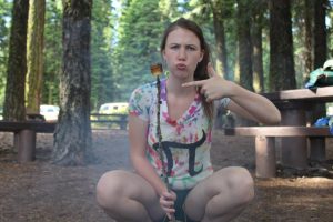A young woman crouches outdoors in a wooded area, holding a stick with a roasted marshmallow and pointing at it while making a playful face. Picnic tables and trees are visible in the background.