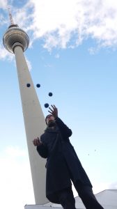 A person in a dark coat is juggling blue balls in front of a tall tower under a bright, partly cloudy sky. The photo is taken from a low angle, emphasizing the height of the tower and the sky above.
