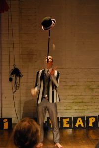 A performer in a striped jacket balances a black hat on a stick held in their mouth, while standing barefoot on a wooden floor in front of a brick wall with painted signs.