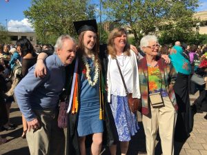 A young woman in a graduation cap and gown stands smiling with three older adults, all with arms around each other, outdoors on a sunny day among a crowd of people.