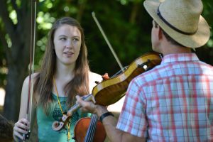 A young woman holding a violin stands outdoors, looking at a man in a straw hat and plaid shirt who is playing a violin. Green trees form a blurred background.