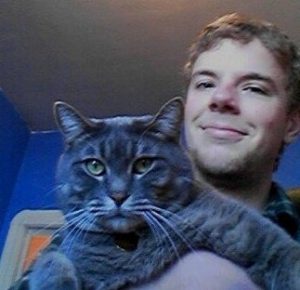 A smiling person holds a large gray tabby cat close to the camera. They are indoors, with blue walls and a beige door visible in the background.