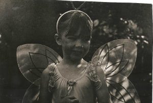 A young child dressed as a fairy with delicate wings and a headband stands outdoors, smiling. The photo is in black and white, with dappled light and a soft background of trees.