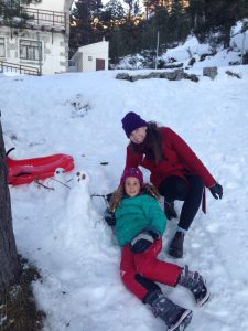 A woman in a red coat and a young girl in winter clothes pose next to a small snowman on a snowy hillside. A red sled is on the ground nearby, and trees and a building are in the background.
